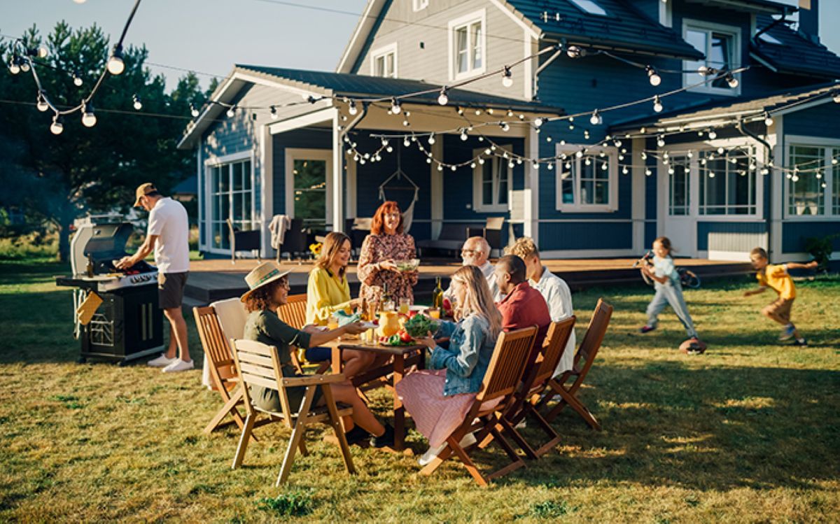 Backyard friends and family barbecue with man grilling, kids playing, and dark gray house in the background