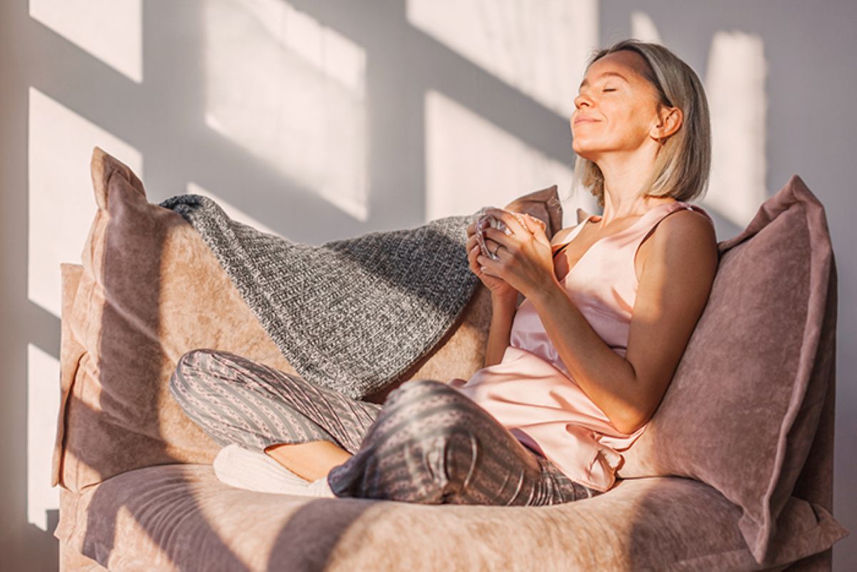 Woman holding cup of coffee and sitting in sunlight on chair inside